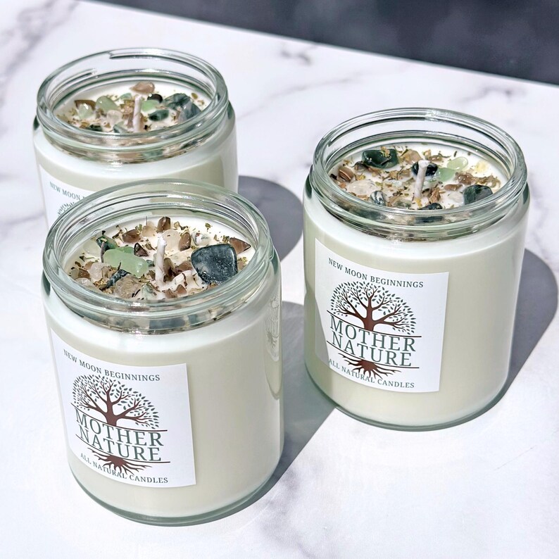 May include: Three glass candle jars with white labels that say "New Moon Beginnings" and "Mother Nature All Natural Candles". The jars are filled with white wax and have green and brown crystals embedded in the wax.