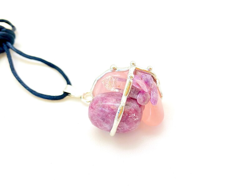 May include: A silver wire cage pendant necklace with pink and purple gemstones. The necklace is made with a black cord.