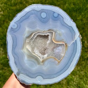May include: A polished, round agate slice with concentric bands of light blue, gray, and beige. The center features a crystalline cavity. The stone is held against a backdrop of green grass, showcasing its natural beauty and intricate patterns.