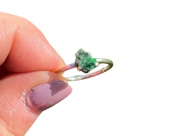 May include: A silver ring with a green gemstone set in the centre. The gemstone is a rough, natural emerald.