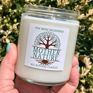May include: A clear glass jar candle with a white label that says "New Moon Beginnings" and features a green tree with brown roots and the text "Mother Nature All Natural Candles".