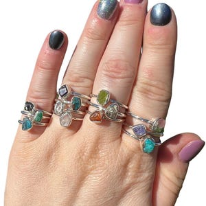 May include: A hand wearing multiple silver rings with different coloured gemstones. The rings are stacked on each finger. The gemstones are small and irregularly shaped.