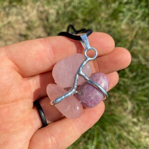 May include: A silver wire wrapped pendant featuring two tumbled stones: a pink rose quartz and a purple lepidolite. The pendant is attached to a black cord.