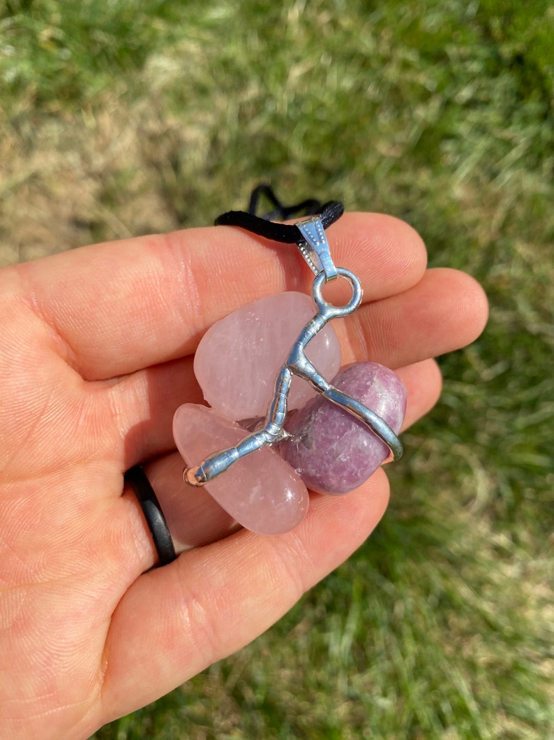 May include: A silver wire wrapped pendant featuring two tumbled stones: a pink rose quartz and a purple lepidolite. The pendant is attached to a black cord.