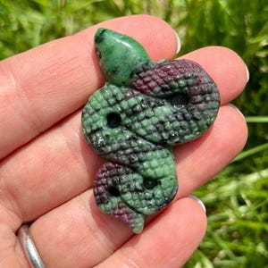 May include: A carved gemstone snake pendant. The snake is green and burgundy with a detailed scale pattern. The snake's head is a glossy green, and the body is a mix of green and burgundy. The pendant is held in a hand against a background of green grass.