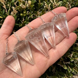 May include: Four clear quartz crystal pendulums of varying sizes with silver chains. The pendulums are arranged in a hand, with the largest pendulum at the top and the smallest pendulum at the bottom.