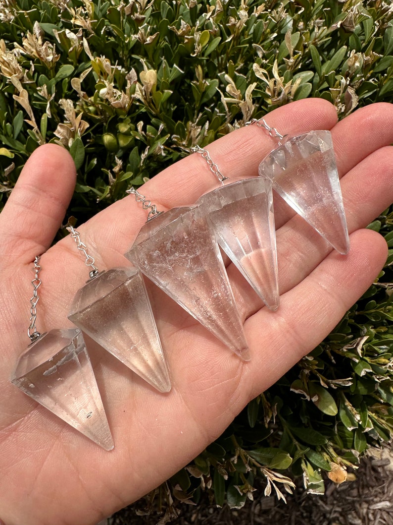May include: Four clear quartz crystal pendulums of varying sizes with silver chains. The pendulums are arranged in a hand, with the largest pendulum at the top and the smallest pendulum at the bottom.