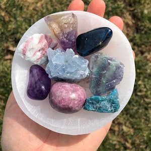Pu&ograve; includere: Una collezione di otto cristalli di vari colori, tra cui viola, rosa, blu, verde e marrone, sono esposti in una ciotola di selenite bianca. I cristalli sono disposti in un motivo circolare.