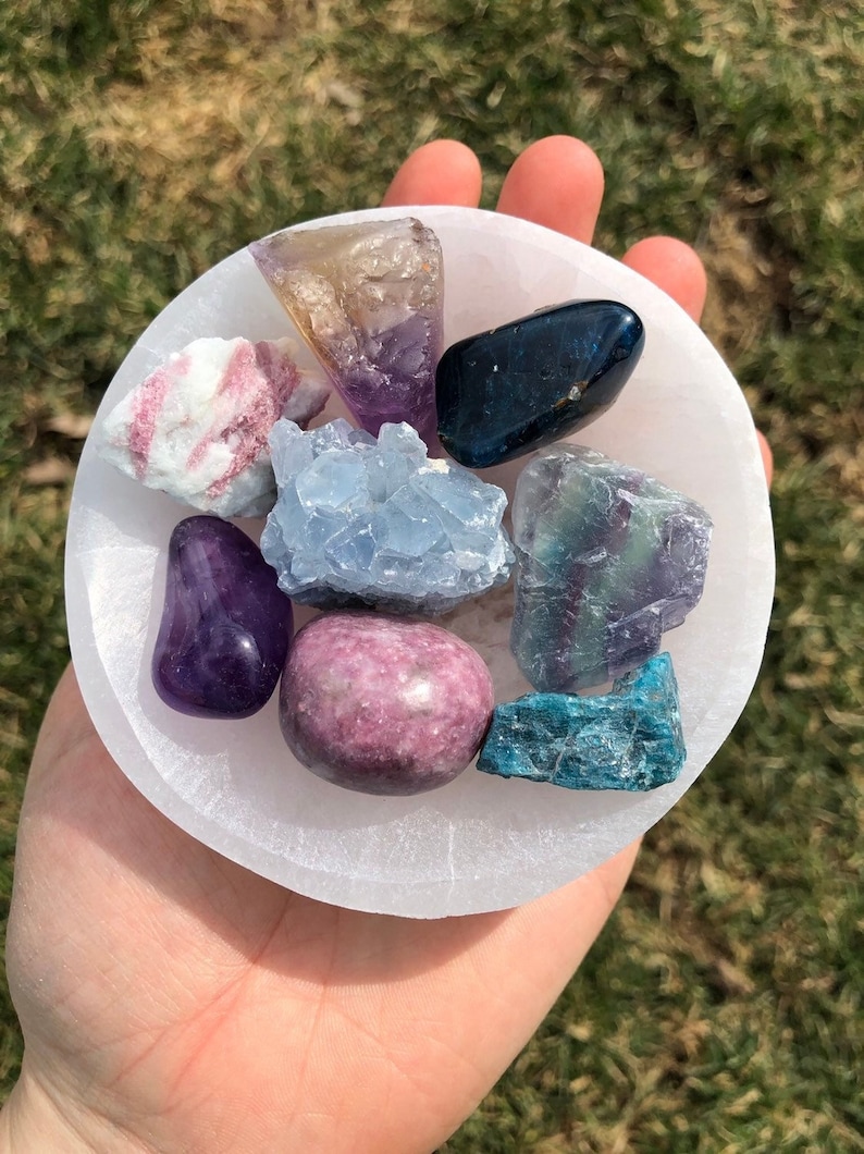 Pu&ograve; includere: Una collezione di otto cristalli di vari colori, tra cui viola, rosa, blu, verde e marrone, sono esposti in una ciotola di selenite bianca. I cristalli sono disposti in un motivo circolare.