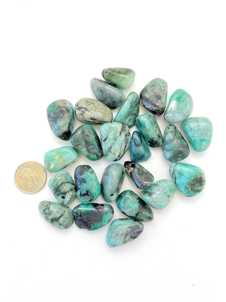 Puede incluir: Una colecci&oacute;n de piedras de esmeralda pulidas, que var&iacute;an en tama&ntilde;o y tono de verde. Las piedras tienen una textura natural y rugosa y est&aacute;n dispuestas sobre un fondo blanco. Se incluye una moneda de un centavo de d&oacute;lar estadounidense para la escala.