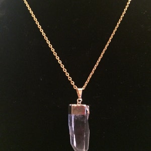 Puede incluir: Un collar de cadena dorada con un colgante de cristal de cuarzo transparente. El colgante es un cristal largo y puntiagudo con una tapa dorada en la parte superior.