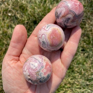 May include: Three pink and grey patterned spheres of polished pink calcite, also known as  pink aragonite, in a hand.