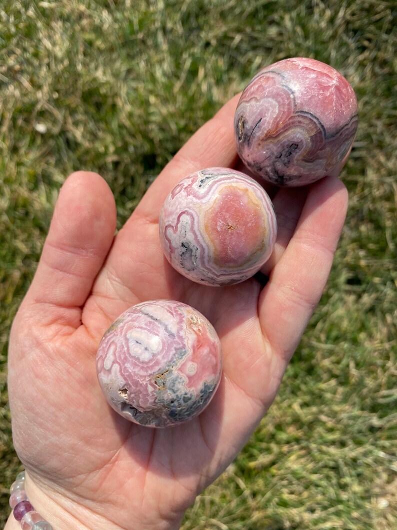 May include: Three pink and grey patterned spheres of polished pink calcite, also known as  pink aragonite, in a hand.