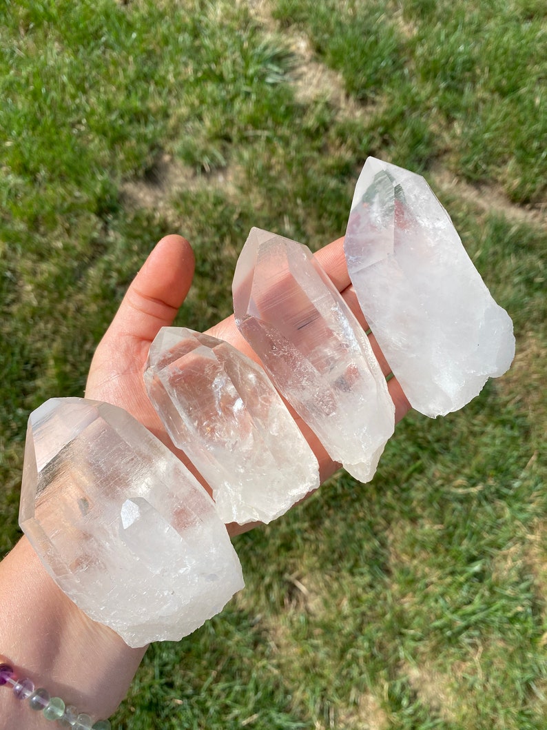 May include: Four clear quartz crystal points, also known as rock crystals, are arranged in a row on a hand. The crystals are various sizes and shapes, with some having a more pointed tip than others.