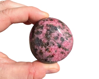 May include: A smooth, polished pink and black gemstone with a mottled pattern. The stone is shaped like an oval and is being held in a hand.