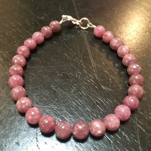 K&ouml;nnte beinhalten: Ein Armband aus rosafarbenem Lepidolith mit einem silbernen Verschluss. Das Armband besteht aus runden Perlen und ist etwa 18 cm lang.