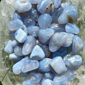 May include: A collection of polished blue lace agate stones. The stones vary in size and shape, with a mix of light and darker blue hues. Some stones have white and brown inclusions, and they are displayed in a clear bowl.