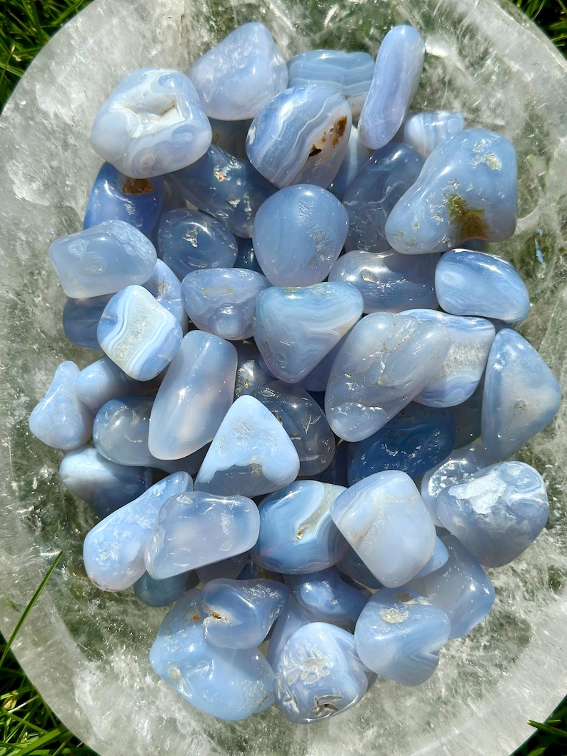 May include: A collection of polished blue lace agate stones. The stones vary in size and shape, with a mix of light and darker blue hues. Some stones have white and brown inclusions, and they are displayed in a clear bowl.