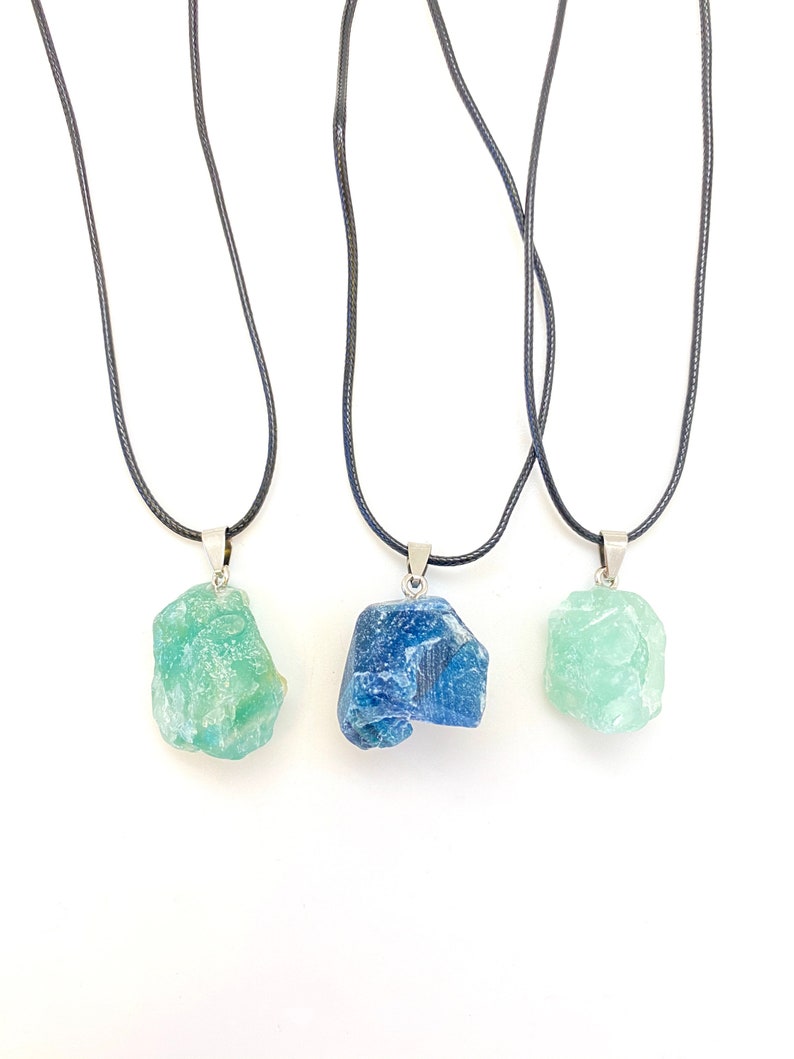 Puede incluir: Tres collares con colgantes de cristal en bruto. Los colgantes son verdes, azules y verdes. Los collares est&aacute;n sobre un fondo blanco.