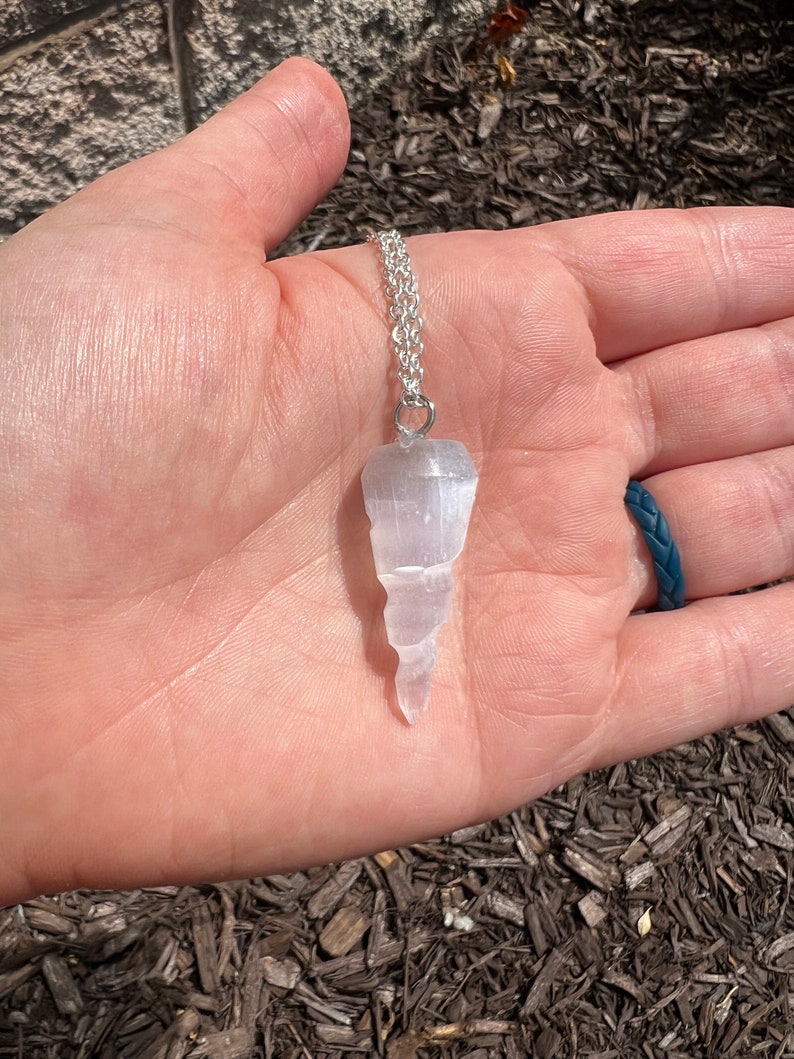 K&ouml;nnte beinhalten: Ein wei&szlig;er Selenit-Kristall-Anh&auml;nger an einer silbernen Kette. Der Kristall ist in Form eines spitzen Eiszapfens geformt und hat eine glatte, polierte Oberfl&auml;che.