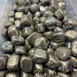 May include: A close-up image of a large pile of tumbled pyrite stones. The stones are a metallic gold color and have a smooth, rounded surface.