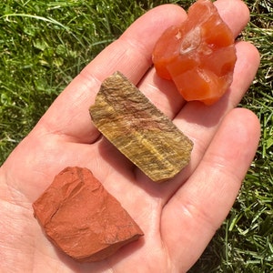 May include: Three rough, natural stones: a red-brown stone, a golden tiger's eye stone, and a bright orange carnelian stone.
