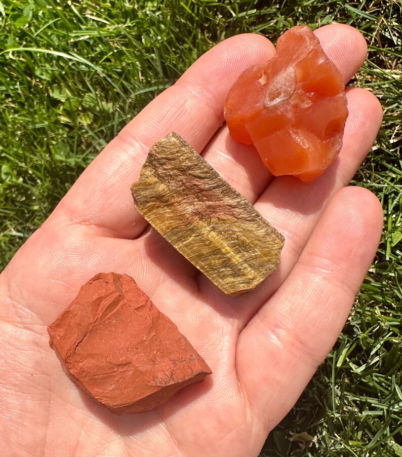 May include: Three rough, natural stones: a red-brown stone, a golden tiger's eye stone, and a bright orange carnelian stone.