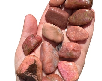 May include: A collection of polished, tumbled stones in shades of pink, red, and brown. The stones vary in size and shape, with smooth, rounded surfaces. Some stones have darker, contrasting patterns.