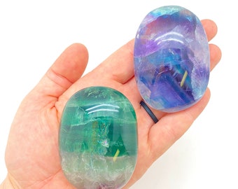 Puede incluir: Dos piedras de palma de fluorita pulidas. Una es verde y morada, la otra azul y morada. Las piedras tienen forma ovalada y se sostienen en una mano sobre un fondo blanco. Las piedras son translúcidas.