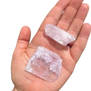 Puede incluir: Dos cristales de color lila claro con apariencia transl&uacute;cida se sostienen en una mano abierta. Los cristales tienen una superficie rugosa y texturizada y formas variables. El fondo es blanco liso.