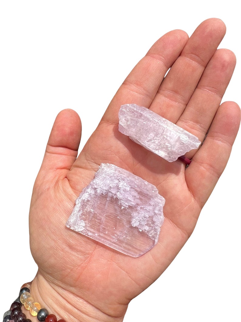 Puede incluir: Dos cristales de color lila claro con apariencia transl&uacute;cida se sostienen en una mano abierta. Los cristales tienen una superficie rugosa y texturizada y formas variables. El fondo es blanco liso.