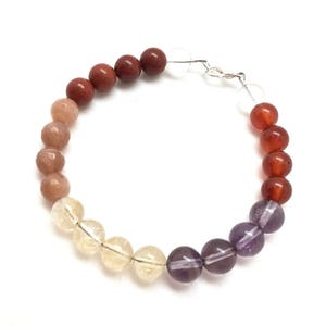 Puede incluir: Un brazalete de piedras preciosas multicolores con cuentas marrones, naranjas, amarillas y moradas. El brazalete está hecho con piedras naturales y tiene un cierre plateado.