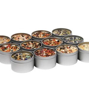 May include: A collection of twelve round silver metal tins containing white wax candles topped with various colorful crushed gemstones and crystals. Each candle features a central wick and a unique arrangement of mineral fragments in shades of red, blue, orange, and clear.