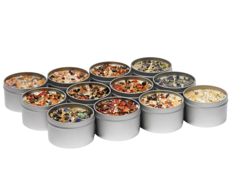 May include: A collection of twelve round silver metal tins containing white wax candles topped with various colorful crushed gemstones and crystals. Each candle features a central wick and a unique arrangement of mineral fragments in shades of red, blue, orange, and clear.