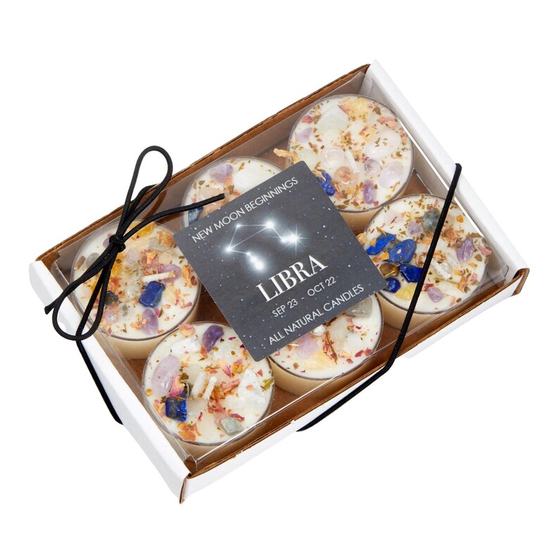 May include: A set of six small candles in a white box with a black ribbon. Each candle is topped with various colorful crystals and dried flower petals. A square black label features the text New Moon Beginnings Libra Sep 23 - Oct 22.