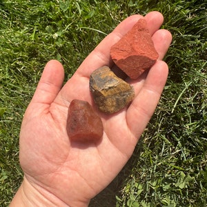 May include: Three tumbled stones in a hand. A red jasper stone, a tiger's eye stone, and a red carnelian stone.