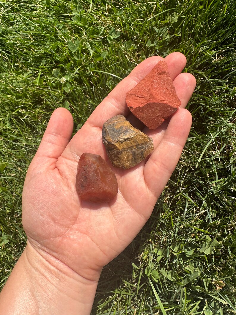 May include: Three tumbled stones in a hand. A red jasper stone, a tiger's eye stone, and a red carnelian stone.