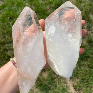 May include: Two clear quartz crystal points, one is larger than the other, with a slightly cloudy appearance. The crystals are held in a hand against a green grassy background.