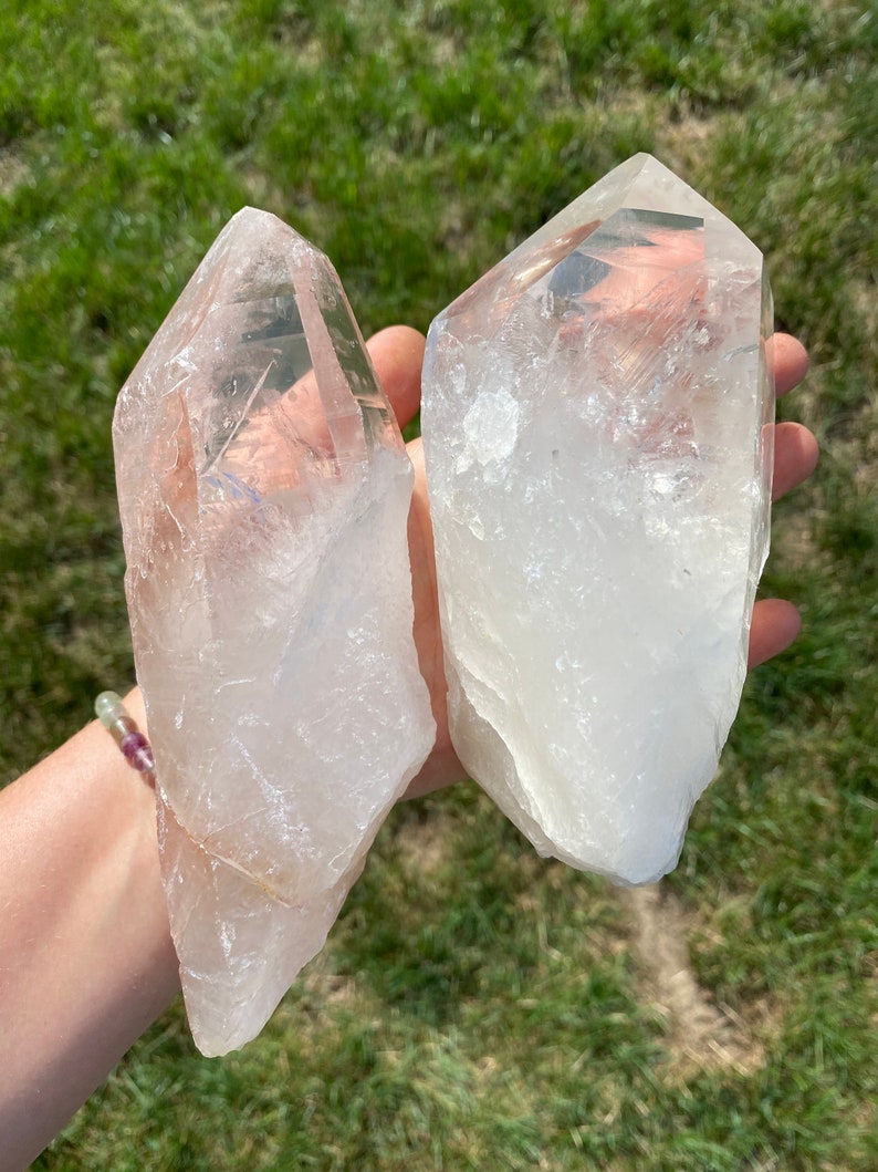 May include: Two clear quartz crystal points, one is larger than the other, with a slightly cloudy appearance. The crystals are held in a hand against a green grassy background.