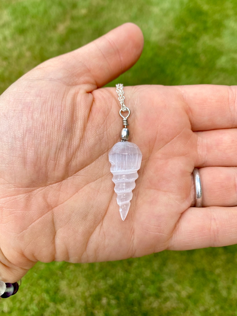K&ouml;nnte beinhalten: Ein wei&szlig;er Selenit-Kristall-Anh&auml;nger in Form einer Spirale, der an einer silbernen Kette h&auml;ngt. Der Anh&auml;nger ist etwa 5 cm lang und wird in einer Hand gehalten.