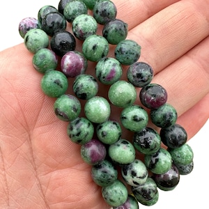 May include: A strand of green, black, and pink gemstone beads. The beads are round and have a speckled appearance.