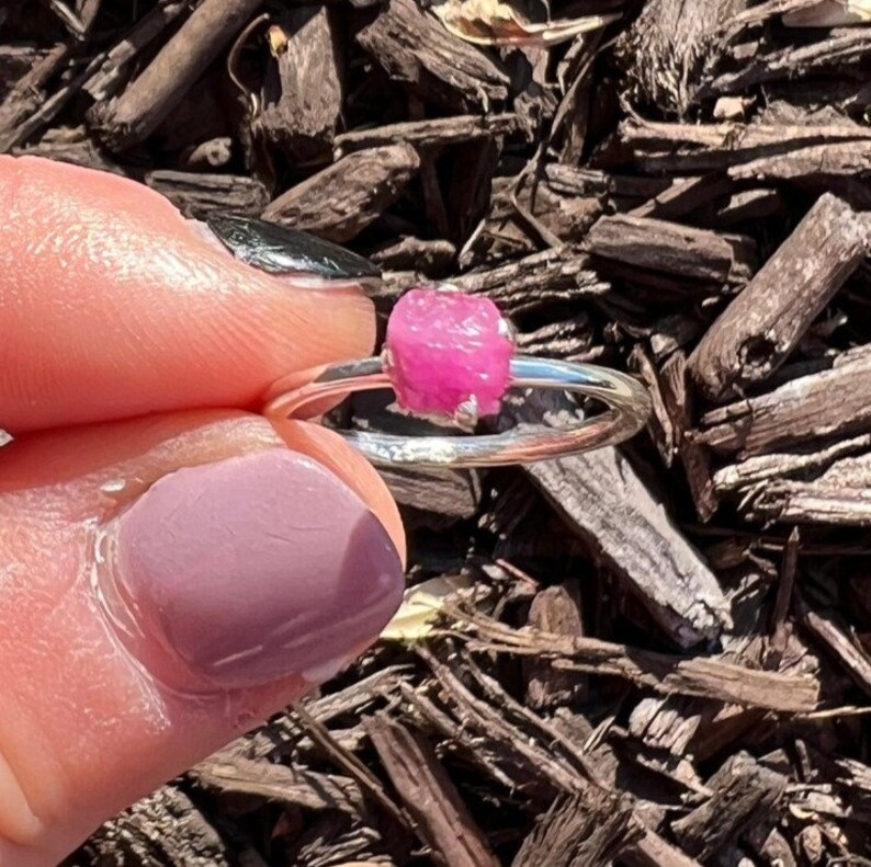 May include: A silver ring with a pink, rough-cut gemstone. The ring is on a finger.