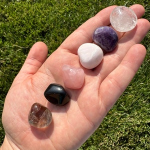 Puede incluir: Una colecci&oacute;n de siete piedras pulidas, incluyendo cuarzo transparente, amatista, cuarzo rosa, cuarzo ahumado y obsidiana negra. Las piedras est&aacute;n dispuestas en una mano, con el cuarzo transparente en la parte superior y la obsidiana negra en la parte inferior.