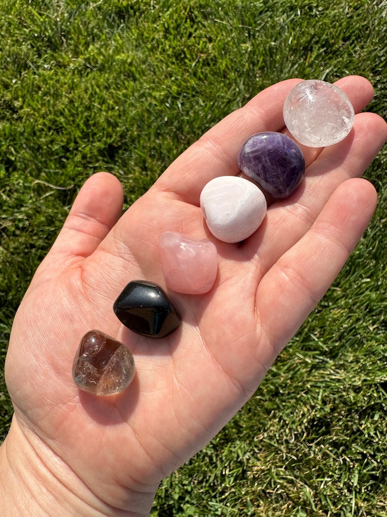 Puede incluir: Una colecci&oacute;n de siete piedras pulidas, incluyendo cuarzo transparente, amatista, cuarzo rosa, cuarzo ahumado y obsidiana negra. Las piedras est&aacute;n dispuestas en una mano, con el cuarzo transparente en la parte superior y la obsidiana negra en la parte inferior.