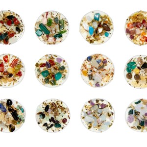 May include: Twelve round white candle holders with various colorful crystals and stones embedded in the wax. The crystals are a mix of clear, green, red, brown, and gold.
