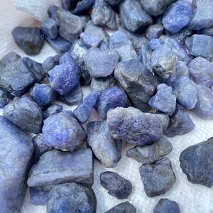 May include: A collection of rough, raw, blue-purple gemstones. The stones are various sizes and shapes, and have a natural, unpolished finish.