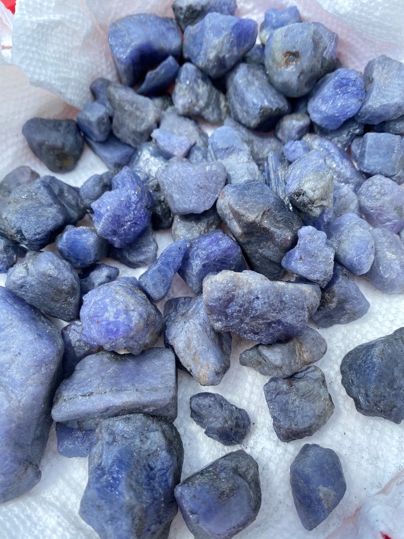 May include: A collection of rough, raw, blue-purple gemstones. The stones are various sizes and shapes, and have a natural, unpolished finish.