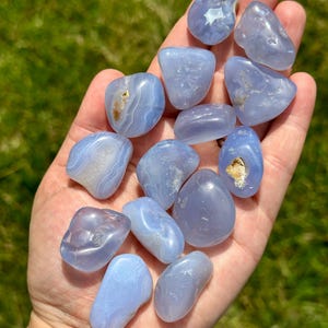 May include: A collection of smooth, polished blue chalcedony stones. The stones vary in shape and size, with some displaying white and brown inclusions. The stones are held in a hand against a backdrop of green grass.