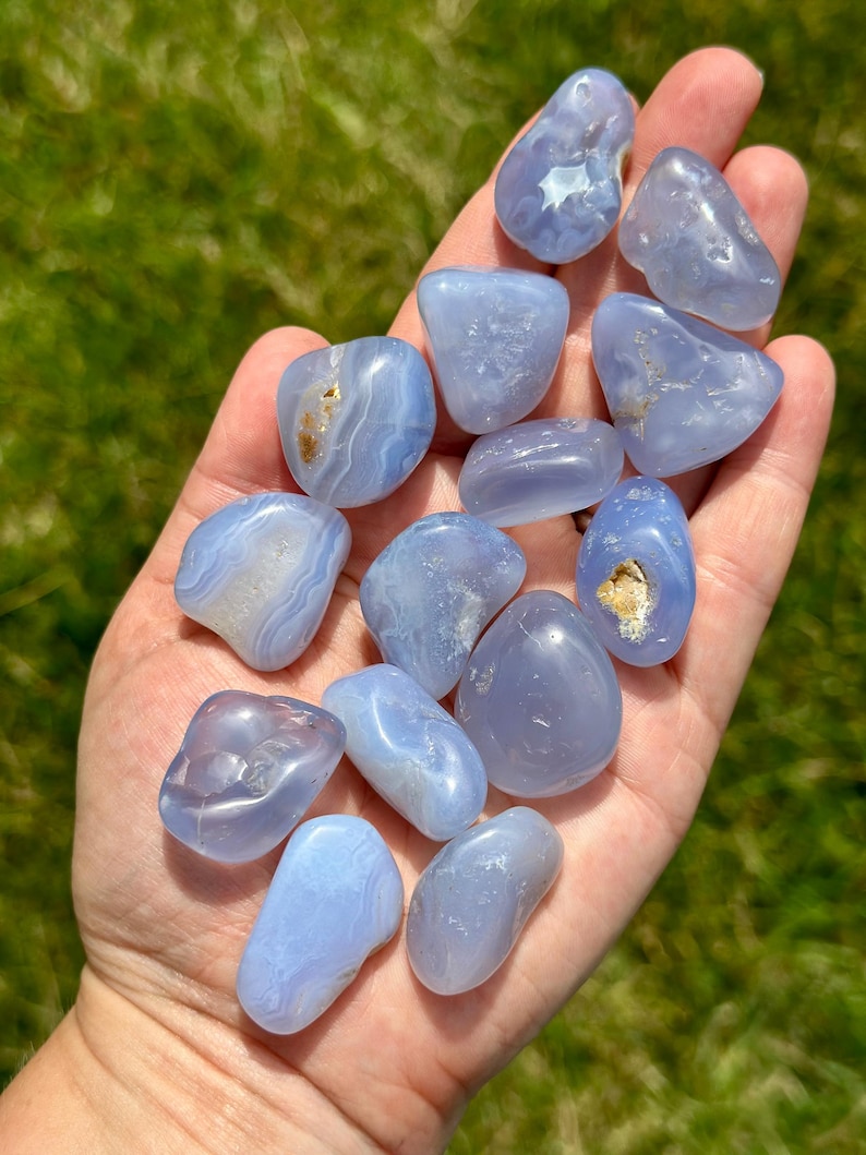May include: A collection of smooth, polished blue chalcedony stones. The stones vary in shape and size, with some displaying white and brown inclusions. The stones are held in a hand against a backdrop of green grass.