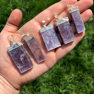 May include: Six purple lepidolite crystal pendants with silver settings. The pendants are rectangular in shape and have a smooth, polished finish.